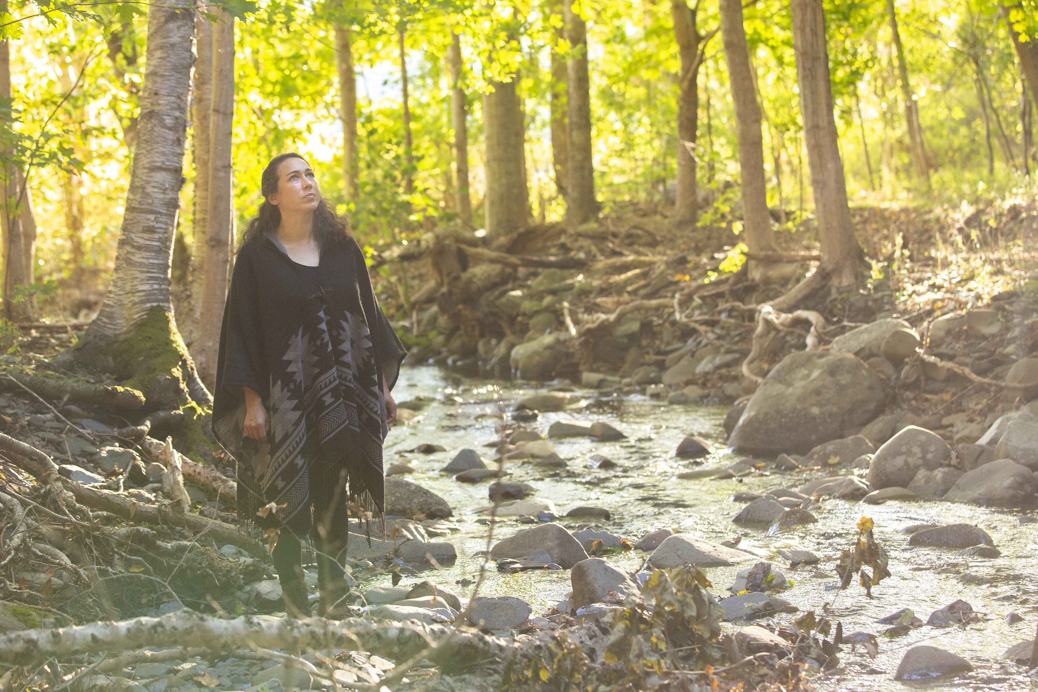 A woman stands in a forest next to a creek.