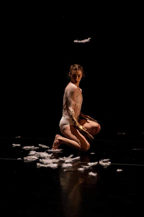 A dancer in a white bodysuit kneels among white feathers.