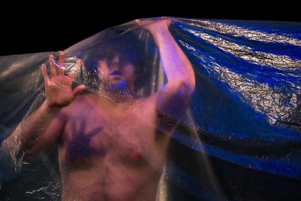 A shirtless man reaches one hand forwards and another lifts at the clear plastic sheet that drapes over his whole body.