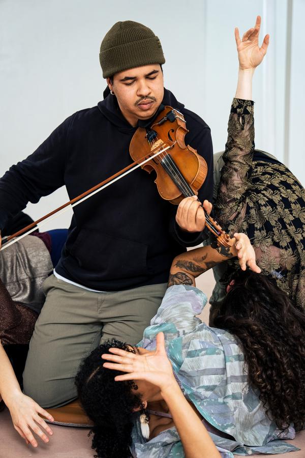 A man kneels playing a violin, surrounded by dancers reaching their hands.