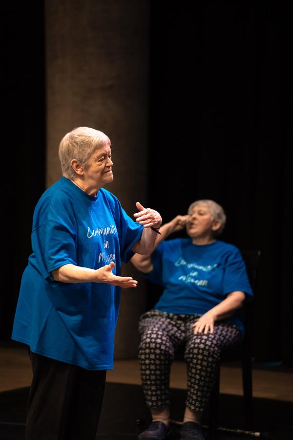 Two elderly women dance, one sitting, one standing.